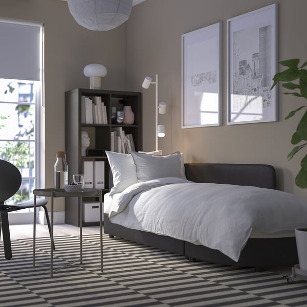 Modern bedroom with sofa bed, white linens, rug, lamp, shelf, art, window.