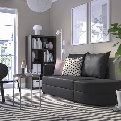 Minimalist living room with grey sofa bed, pillows, black bookshelf, glass table, striped rug, art, balcony.
