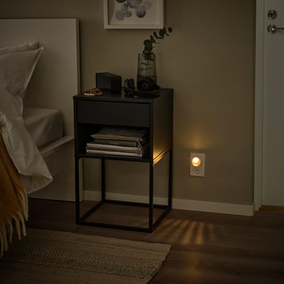 LJUSTER nightlight near bedside table with books, vase, and lamp. Bed, rug on wooden floor.