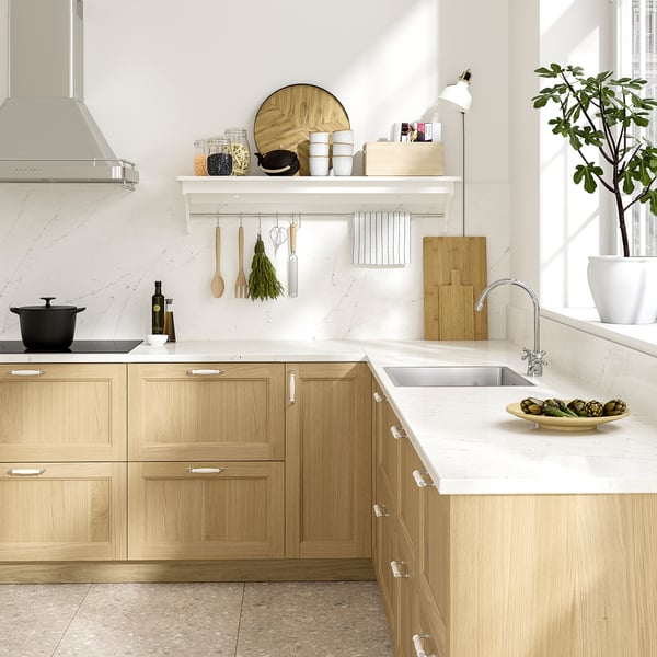 Modern kitchen with light wooden cabinets, white marble countertops, stainless sink, hanging utensils, and a potted plant. Bright and spacious feel.
