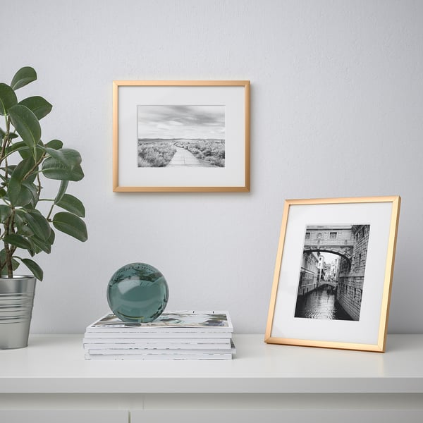 White shelf with gold-framed photos, greenery, and teal sphere. One photo shows a pathway, the other a canal bridge.