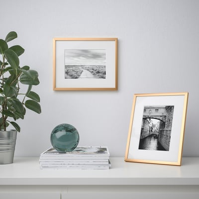 White shelf with gold-framed photos, greenery, and teal sphere. One photo shows a pathway, the other a canal bridge.