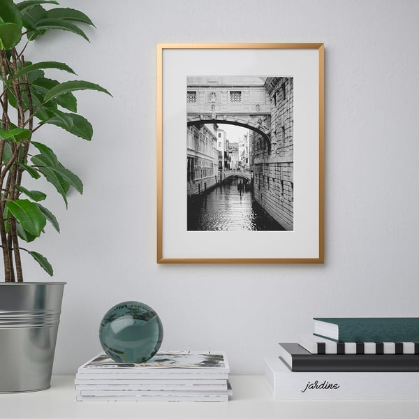 A framed black-and-white a bridge over a canal hangs on a wall above a stack of magazines. A potted plant is to the left.