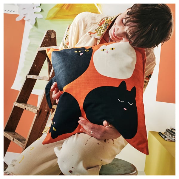 Person holding MÄVINN cat cushion cover, featuring playful cat designs. Cover is black and white cat on red, made of 100% cotton.