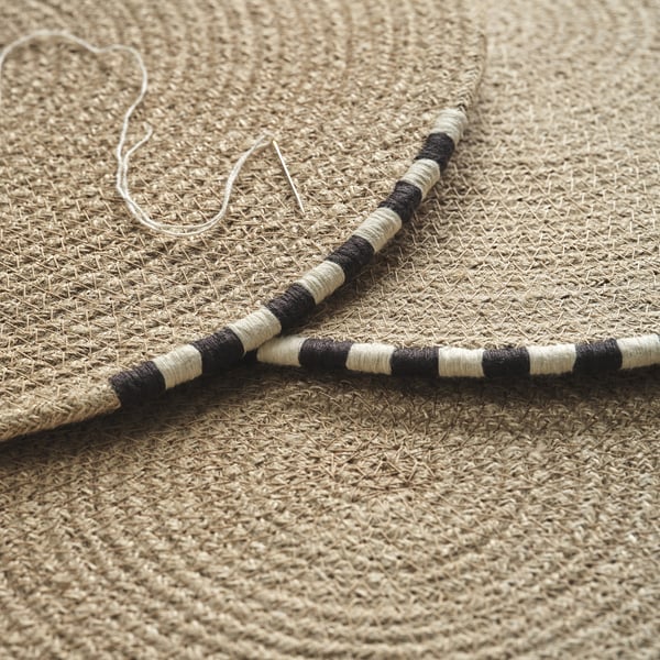 Natural woven jute placemat with hand-stitched black and white edge detail, laid out flat.