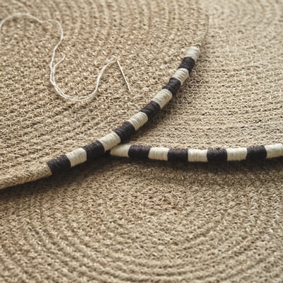Natural woven jute placemat with hand-stitched black and white edge detail, laid out flat.
