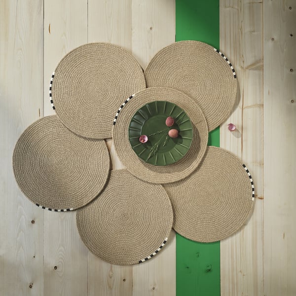 Four woven jute place mats surround a green plate with pink fruits on a wooden floor.