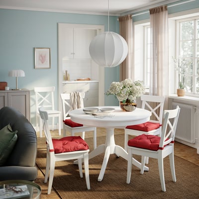 A cosy dining area with a white round table, four white chairs with red cushions, and a pendant light.