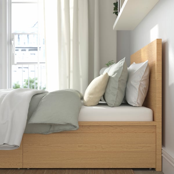 Modern wooden bed with large storage drawers and pillows, near large windows, providing comfort and space.