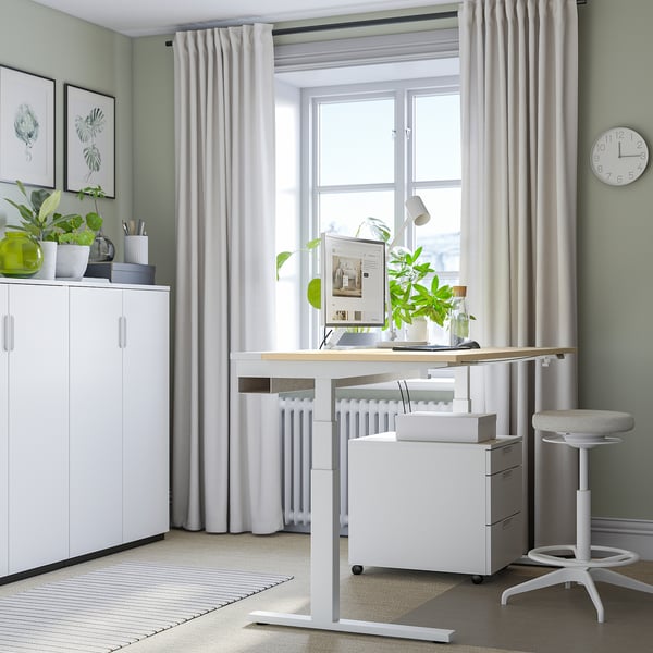 Modern home office setup with white MITTZON standing desk, chair, and storage. Large, bright window, potted plants, and minimalist decor.