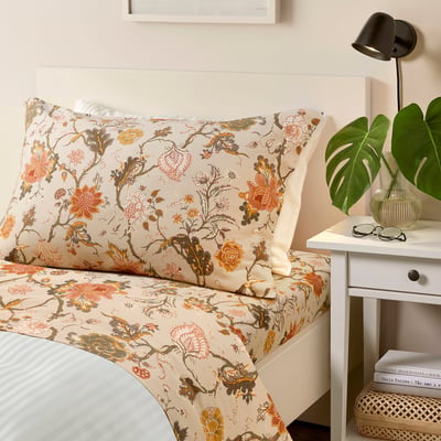 Cosy bedroom with floral bed, beige NÄSSELKLOCKA bedding, white bedside table, black lamp, green plant, and books.