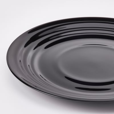 Round, black plate with a glossy, rippled design on a white surface, reflecting light, showing high-quality craftsmanship and sleek finish.