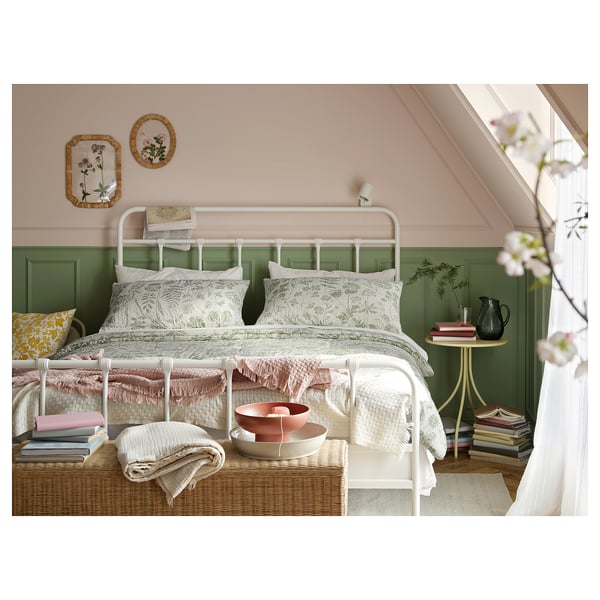 Cosy bedroom with white metal bed, green wall panels, floral bedding, wicker chest, and pastel decorations.