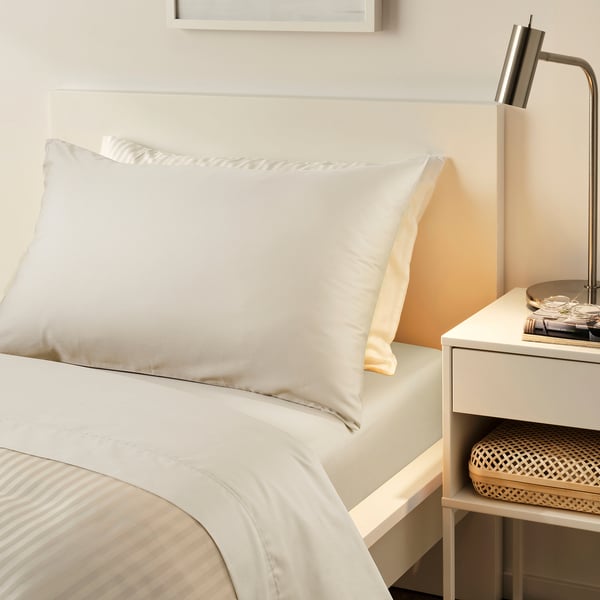 NATTJASMIN cream bed set. Silky, sateen-woven cotton/lyocell. Soft, lustrous pillow and flat sheet atop minimalist bed. Beside compact bedside table.