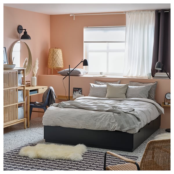 Modern minimalist bedroom NORDLI bed with grey linens and wooden drawers. Light pink walls, large window with sheer curtains, and black desk lamp.