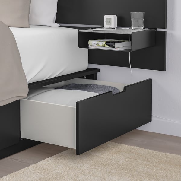 Bedside setup with wall-mounted shelf, alarm clock, glass, books, and partially open under-bed storage drawer.