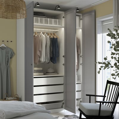 A modern wardrobe with open doors showing clothes and drawers, integrated lighting, and adjustable feet. A chair and dressing gown hang nearby.
