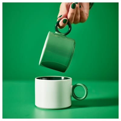 A hand holds a green PIGGÅL mug over a white one. Green mug has a unique round handle, simple design, and colourful glaze.