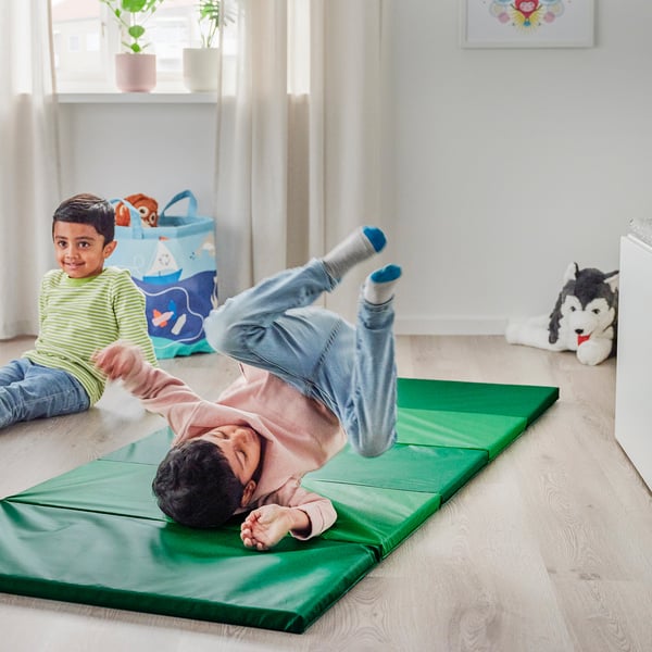 Kids playing on a soft, foldable green exercise mat indoors. One is doing a handstand, the other is sitting nearby, with a stuffed animal close by.