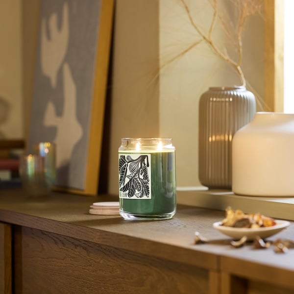 PRAKTHÄGER green candle sits on wood shelf. Near vases, picture, bowl. Its glass, cylindrical with leaf design. Smaller than vases, larger than bowl.