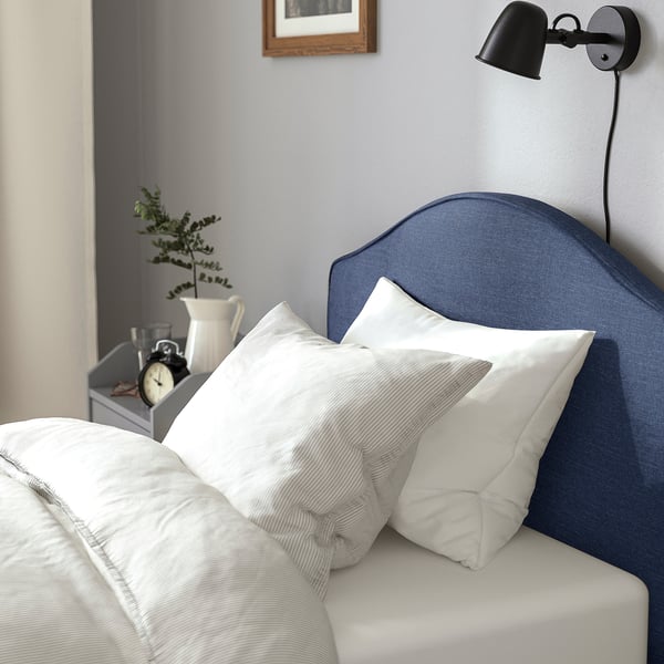 A cosy bed with a padded headboard. Nearby, a wall lamp illuminates a bedside table with a clock, vase, and plant.