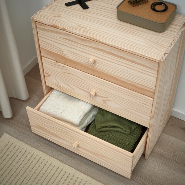 Light wooden dresser with one open drawer revealing white and green sweater, positioned against wall.