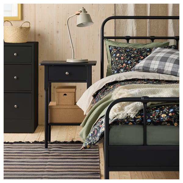 A cosy bedroom with a black metal bed, dark floral bedding, and a striped rug. A wooden bedside table with a lamp and storage boxes is also visible.