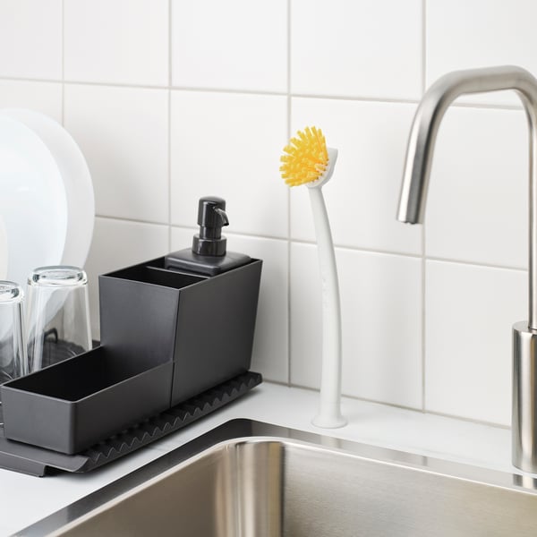A kitchen sink area with a grey soap dispenser, a yellow-bristled brush with a white handle attached to the wall, and a stainless steel tap.