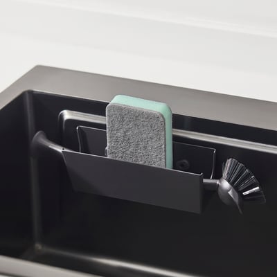 Black sink caddy with green sponge and scrub brush, showing organised storage near sink.