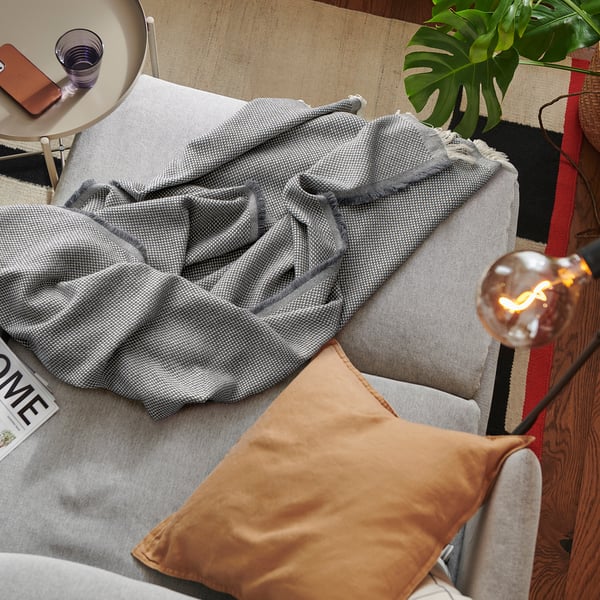 Cosy grey RÖNNBÄRSMAL throw on sofa, next to large orange cushion, lit bulb lamp, and plants.