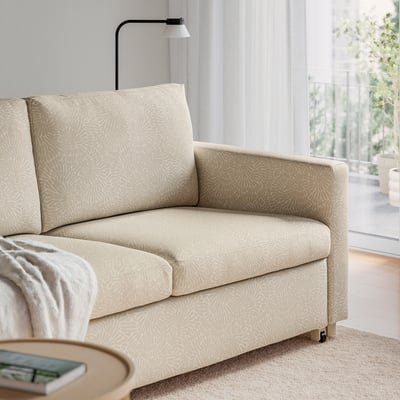 Beige sofa-bed with patterned cover, folded out, near curtained window and small table with book.