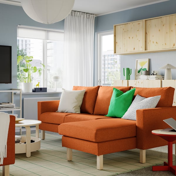Bright living room with orange SALTSJÖBADEN 3-seat sofa w/chaise, green/white pillows, large windows, wooden cabinets, modern decor.