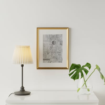 Framed poster on white wall with gold frame. Table below has black lamp with beige shade and green plant in clear vase.