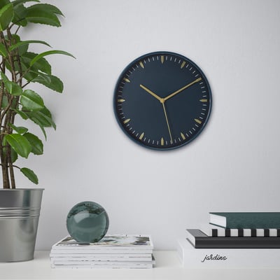 Wall clock with dark blue face and gold hands, silent quartz movement, above table with books and plant.