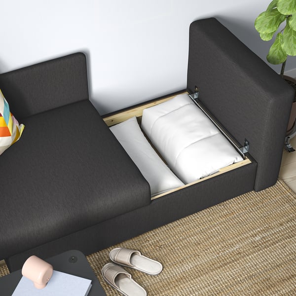 Modern black sofa bed with open storage and folded mattresses, beside a coffee table with lamp and slippers on a rug.