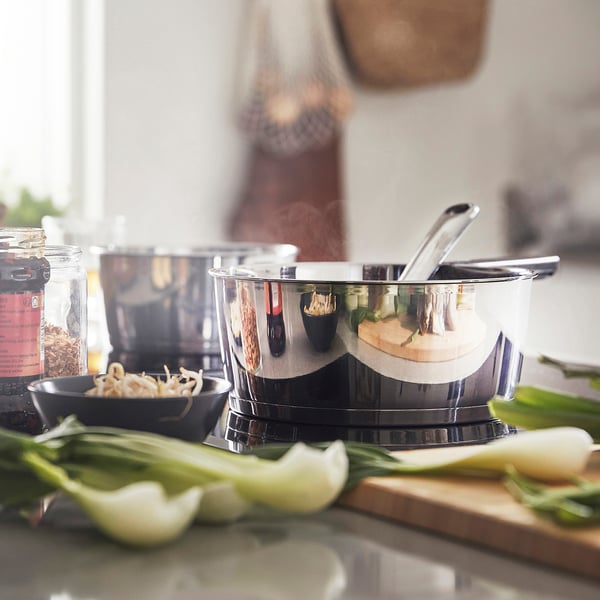 Shiny saucepan with removable handle sits on stove, surrounded by fresh vegetables, ready for cooking.