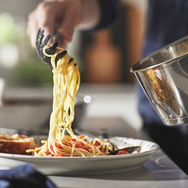 A person lifts a forkful of spaghetti from a plate, showing a detachable handle saucepan nearby, emphasising the products space-saving feature.