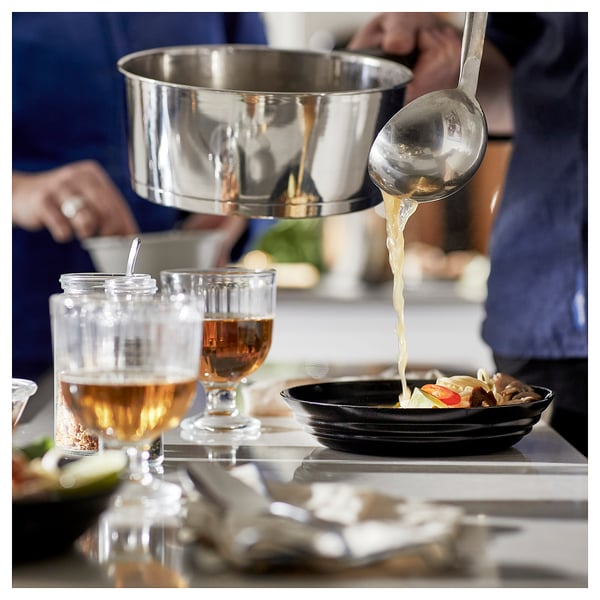 A chef pours sauce from a metal ladle into a black bowl on a counter near wine glasses.