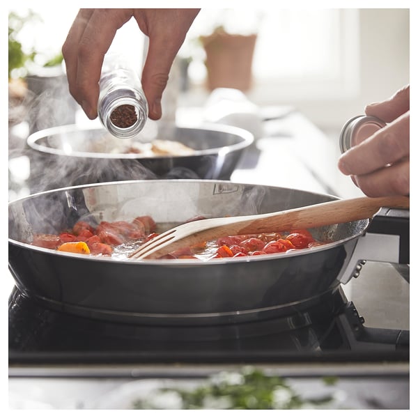 Cooking with SLÄTROCKA pan, adding pepper. Black pan with wood handle, colourful food, steam rising.