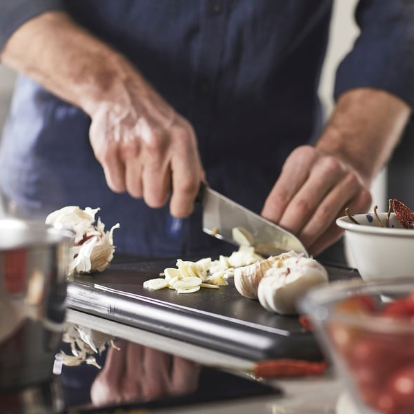 Person chops garlic with a knife on a cutting board. The setup includes a cookware handle, indicating preparation for cooking.