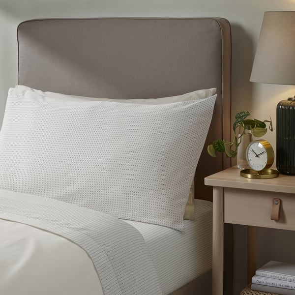 Modern bedroom: grey headboard, white dotted pillow. Alarm clock, plant, lamp on light wood bedside table.