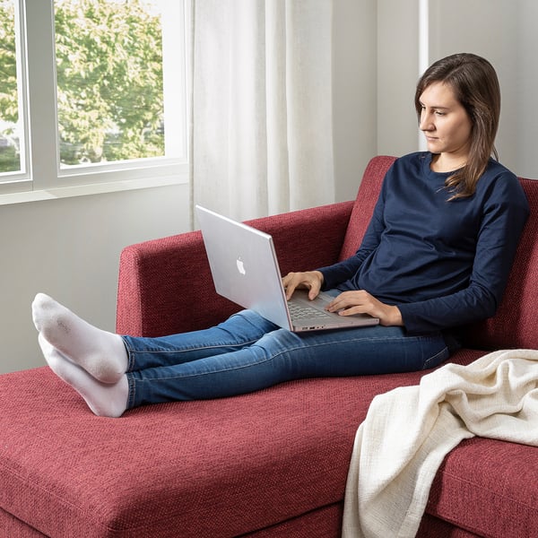Person works on laptop on cosy red couch by window; blanket, comfortable support.