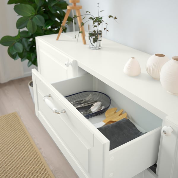 White drawer unit with open drawer revealing utensils and cutting board. Top decorated with plants and vases.