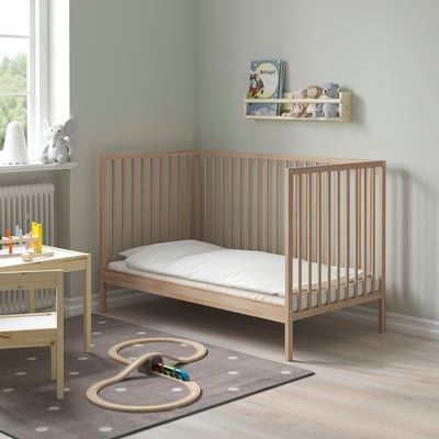 Wooden SNIGLAR cot on polka dot rug, smaller than window, with white mattress and slats for nursery.