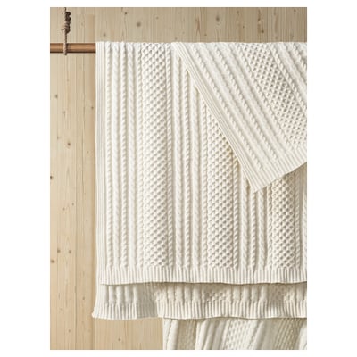 White knitted blanket on wooden wall. Soft, textured, and machine-washable. Made from 100% recycled polyester.