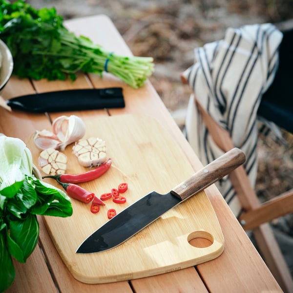 Knife on cutting board with veggies, garlic, herbs. Black blade, wooden handle.
