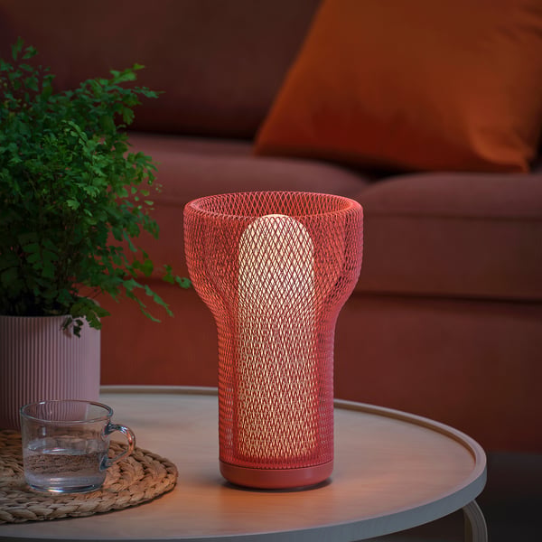 A lit orange table lamp with a mesh design sits on a table next to a plant and a glass cup.