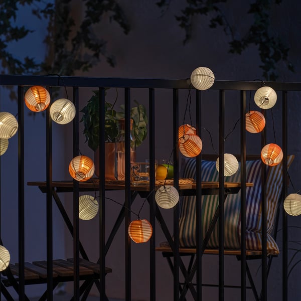Balcony with hanging solar-powered lanterns, creating soft orange and white light. A table holds plants and drinks, with striped chairs visible.