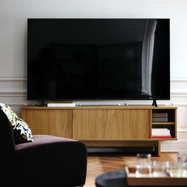 Modern living room with large flat-screen tv on wooden stand, black sofa with patterned pillow, and wooden coffee table with glassware.