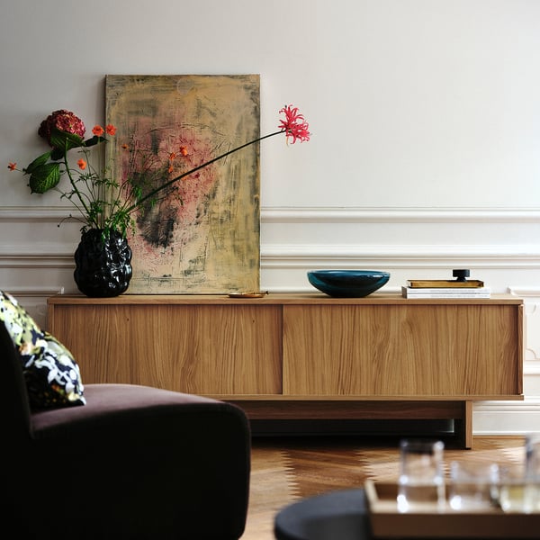 A living room with wooden furniture, an abstract painting, and a black stoneware vase holding colourful flowers on top of a wooden cabinet.